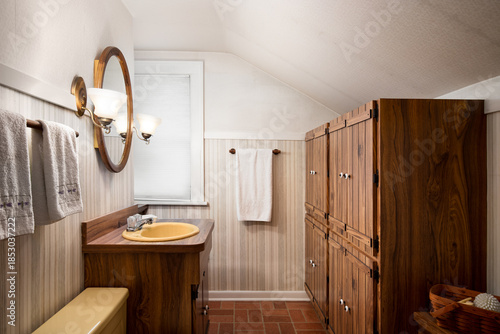 Photos A vintage bathroom featuring warm wood cabinetry, a wooden vanity with a yellow sink, and sconces around the round mirror