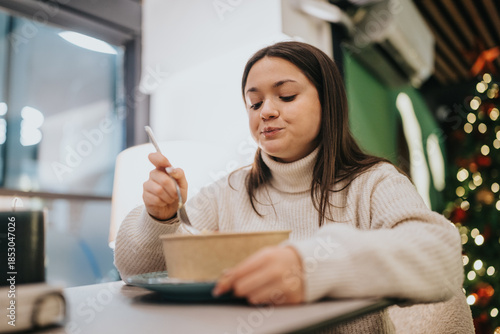 Φωτογραφία A young woman in a cream sweater eats from a bowl at a cafe table