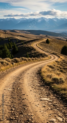 A winding dirt road meanders through rolling hills, with a backdrop of snow-capped mountains under a bright, partly cloudy sky