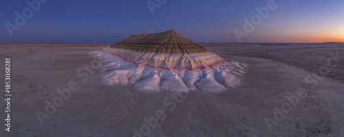 Aerial view of a layered mesa rising from the stark desert floor under a twilight sky, its colors contrasting with the arid landscape, Bokty, Mangystau Region, Kazakhstan.