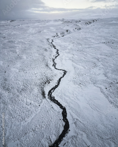 Aerial view of a dark river snaking through a vast, snow-covered landscape under a muted sky, creating a stark contrast between the dark water and the white expanse., Vik, Western Region, Iceland.