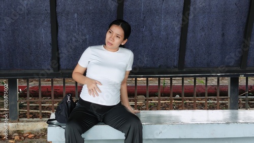 A tired pregnant woman sitting at a modern city bus shelter, looking exhausted while waiting for public transport
