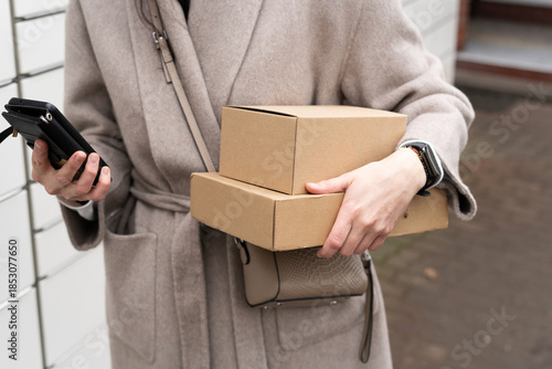Person holding two cardboard boxes and a phone while standing outside a building on a cloudy day