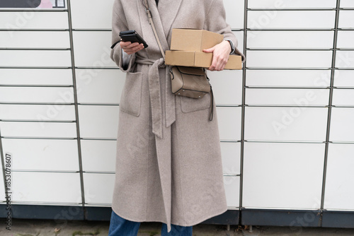 Woman holding boxes and cellphone standing in front of a wall with lockers during the day