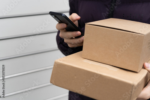 Person holding two delivery boxes and using a smartphone near a wall in a delivery area