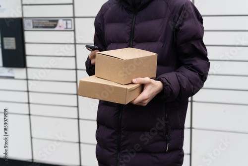 Person holds two cardboard boxes while checking phone near delivery lockers in an urban area