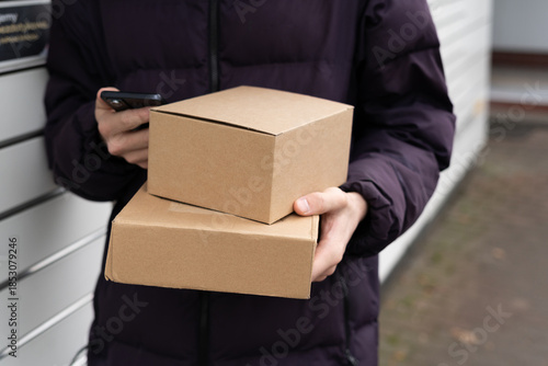Person holding two cardboard boxes while checking a mobile phone outside a building on a cloudy day