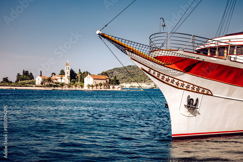 View of the bow of a classic ship with red accents meets the tranquil, shimmering sea, with a distant island and buildings, Vis, Split-Dalmatia County, Croatia.