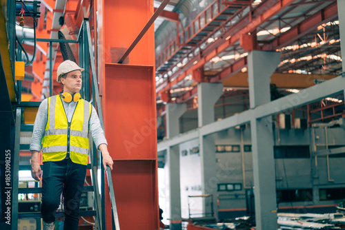 Caucasian industrial engineer man wear safety helmet vest walking down metal stairs in factory. Professional worker holding handrail safety routine. Heavy manufacturing plant background.