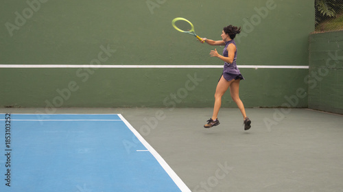 Dynamic action shot of a young woman jumping mid-air to return a tennis shot during practice on an outdoor court. Concept of resilience, high energy, and learning a healthy sports hobby.
