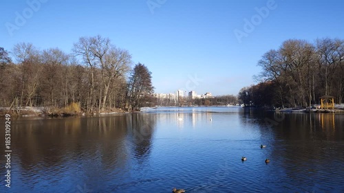 In winter, the lake is covered with ice and snow. There is open water in some places. There is snow on the far shore, the trees of the park are growing and there are buildings. Frost, bright sun
