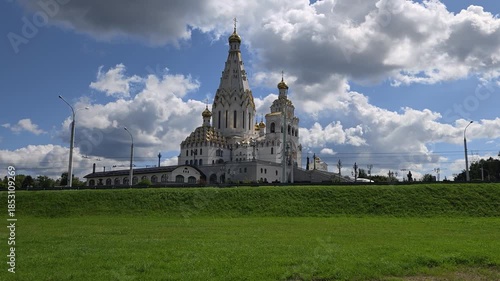 On a grassy lawn stands an Orthodox church with towers, gilded domes and crosses. Nearby there is a roadway of a city street, trolleybus wires and lighting poles. Sunny summer weather and blue sky