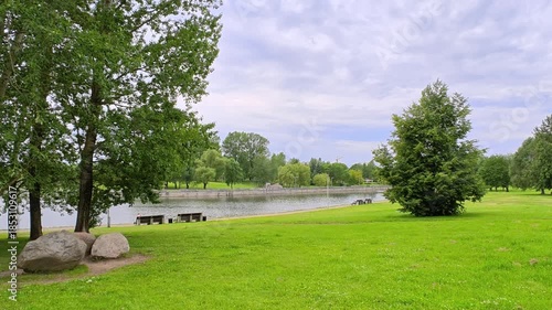 The river flows through the city among grassy lawns with aspens, willows and lime trees growing on them. There are boulders. Fences, benches and lampposts are installed on the concreted shores. Cloudy