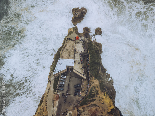 Aerial view of the Fortaleza de Nazaré perched dramatically on a cliff, engulfed by the turbulent Atlantic waves, a striking contrast of earth and sea, Nazaré, Leiria District, Portugal.