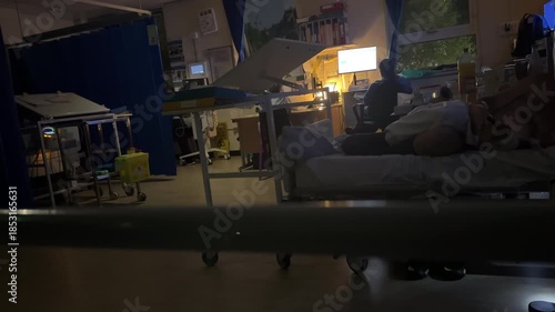 Quiet at night in the dark hospital ward, rear view of two nurses at their desk and computer, patient lying in his bed using mobile phone.