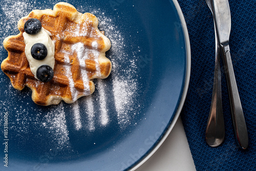 Belgian waffles or dessert with white cream and blueberries sprinkled with powdered sugar on a blue plate with napkin and cutlery. Top view. High quality photo