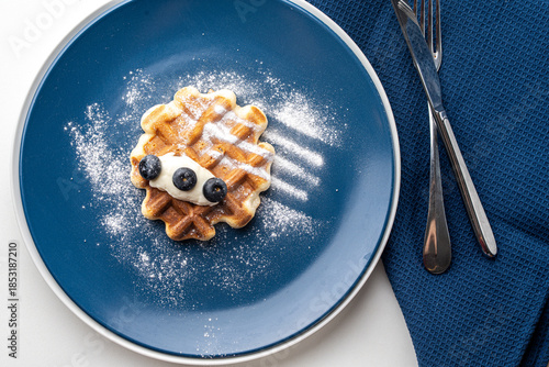 Belgian waffles or dessert with white cream and blueberries sprinkled with powdered sugar on a blue plate with napkin and cutlery. Top view. High quality photo