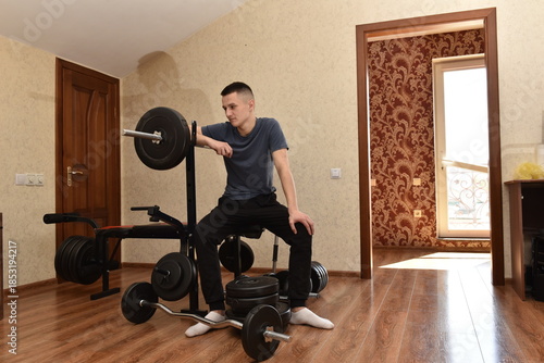 Young man sitting on home gym bench with dumbbells, resting between workouts indoors, casual training at apartment interior with fitness equipment