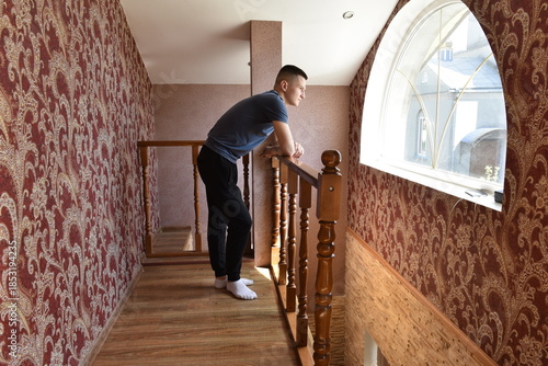 oung man standing on indoor balcony corridor, leaning on wooden railing and looking through window in apartment hallway with patterned wallpaper