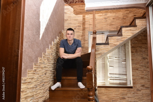 oung man sitting on wooden stairs indoors with crossed arms, looking at camera in modern home interior with stone wall and window light