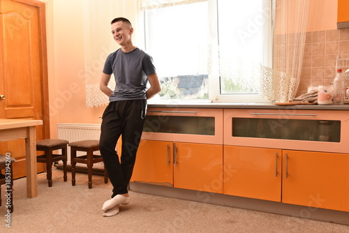 oung man standing barefoot in home kitchen interior, smiling casually near window with natural light, relaxed posture, everyday lifestyle moment