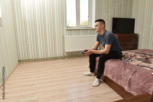 Young man sitting on bed in bright bedroom holding game controller, relaxed at home, casual lifestyle and leisure time indoors
