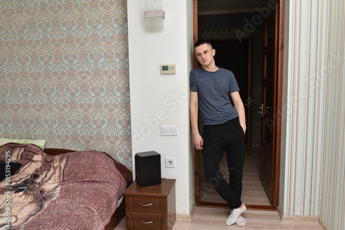 Young man standing in doorway at home, relaxed casual pose in bedroom interior, calm everyday lifestyle moment with soft natural light