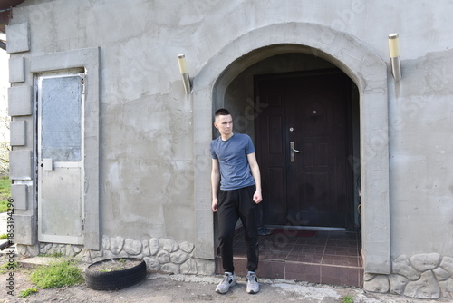 Young man standing at house entrance outdoors, casual clothing, quiet everyday moment near home doorway, simple lifestyle scene in residential exterior