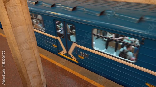Minsk, Belarus - July 13, 2024: Top-down view of a blue subway train with passengers visible through windows, accelerating and leaving platform of marble-columned underground metro station in the city