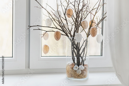 Creative Easter home decor with beige pastel eggs and tree branches in glass vase on windowsill. Bright spring atmosphere, natural light. Home interior.