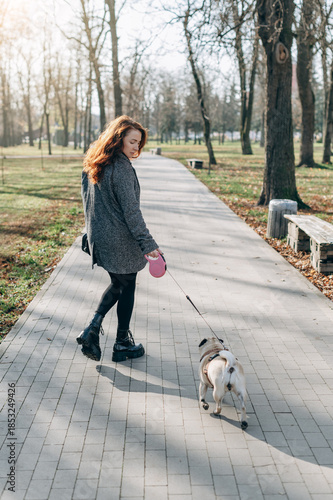 A young woman walks a pug dog on a leash in a public park on a sunny day.