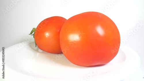 A close-up shot of two ripe, round, vibrant orange-red tomatoes, one with a green stem, slowly spinning on a clean white plate, ideal for food, cooking, health, and market themes.