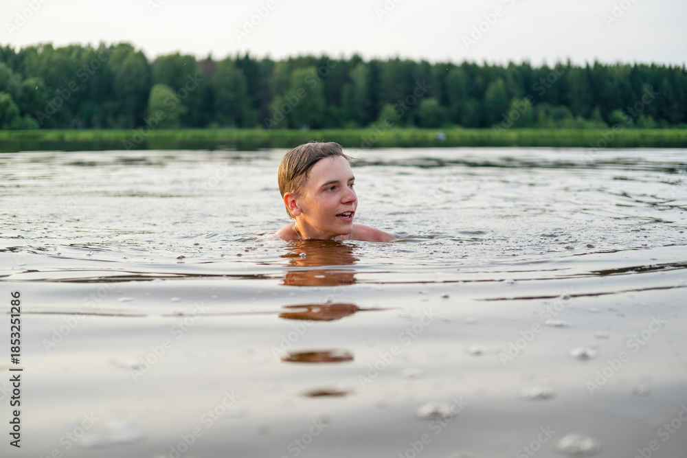 Fototapeta premium A young boy enjoying a swim in a calm lake, surrounded by lush green trees. His expression reflects joy and relaxation as he engages with nature during a peaceful, sunny day.
