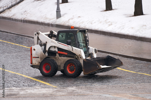 A miniature snow removal tractor with cleared snow in its bucket on the roadway of a city street.