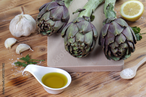 Fresh Artichokes on Cutting Board with Mediterranean Cooking Ingredients: Lemon, Garlic, and Olive Oil on Rustic Wooden Table