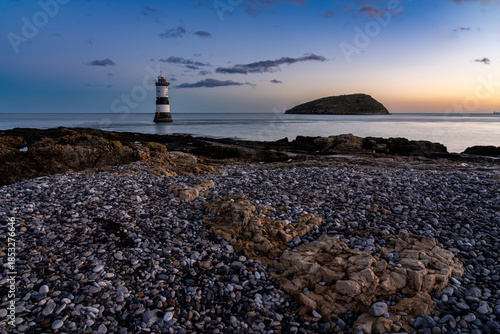 Trwyn Du Lighthouse at Penmon Point at sunrise with calm water and a pebble shoreline in the foreground