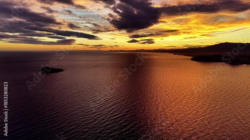 fiery ocean sunset over islet, intense orange sky and dramatic clouds reflect across rippling water, horizon