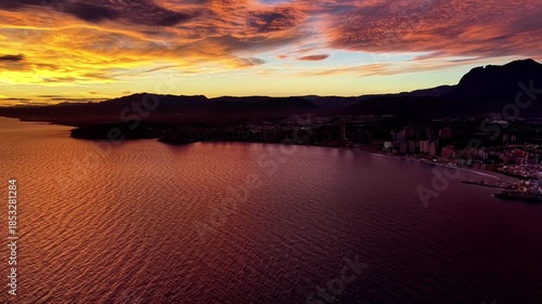 crimson sunset over resort coastline, aerial sweep reveals glowing buildings and calm red sea, evening clouds