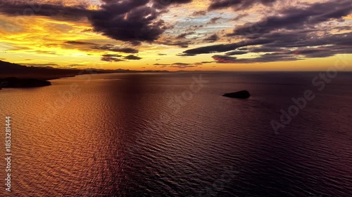 fiery horizon behind solitary rock, drone captures tranquil orange water and textured clouds, intimate coastal