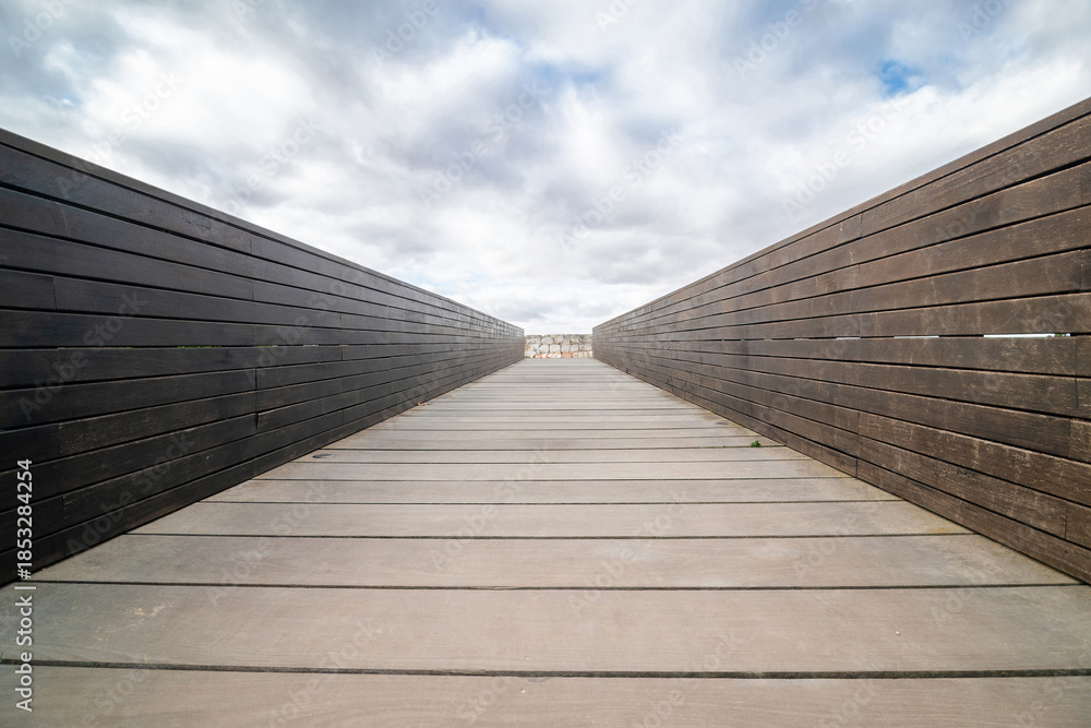 Fototapeta premium Empty wooden walkway under a cloudy sky with a stone wall in the background.