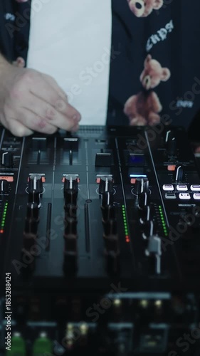 Close up of DJ hands adjusting mixer controls during an energetic electronic music performance with illuminated console buttons and sliders in a dark club setting