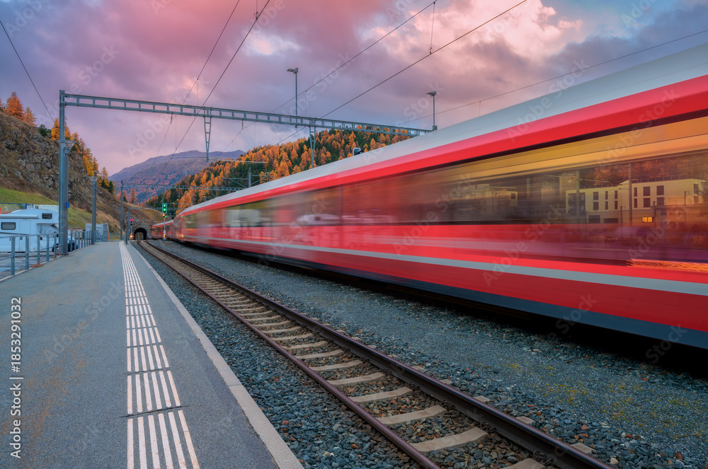 Obraz premium Blurred red passenger train passing mountain railway station in Swiss Alps at dusk. Moving high-speed train. St. Moritz, Switzerland. Bernina Express. Railway platform and cloudy sky. Rail transport