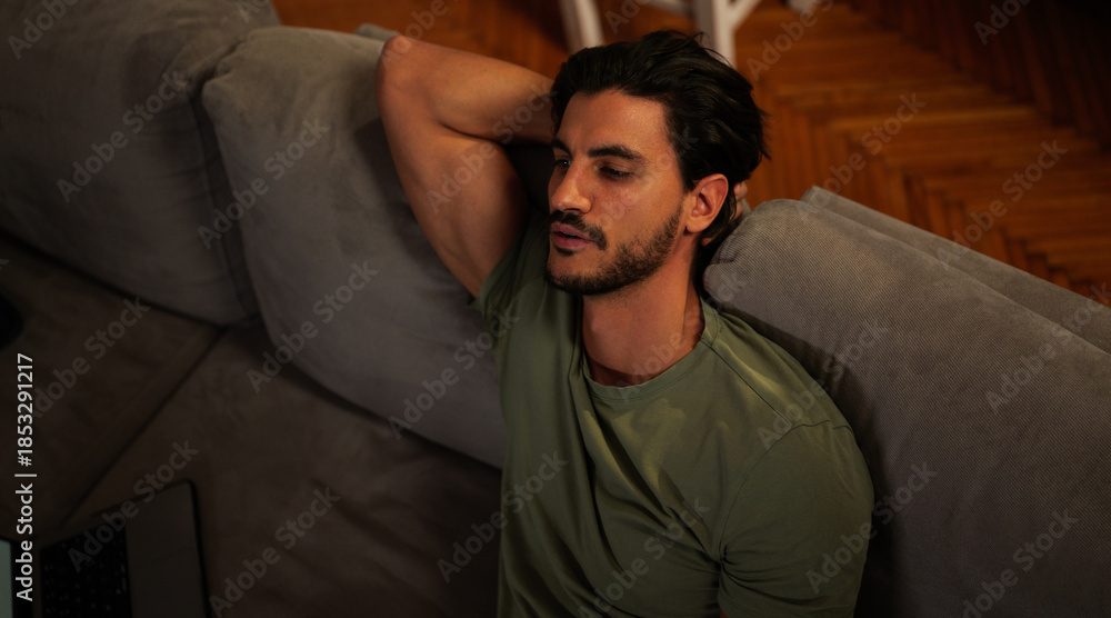 Obraz premium Young man relaxing comfortably on a gray sofa in his living room, casually watching television or a screen, unwinding in a comfortable atmosphere, enjoying leisure time