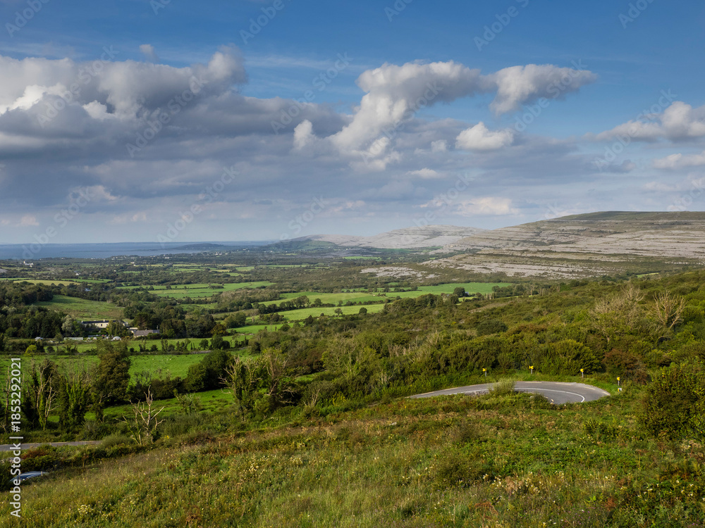 Fototapeta premium Landscape scene with green fields, hills, ocean and blue sky. Burren area, county Galway, Ireland. Warm sunny day. Rural Irish scenery in unique location. Travel and tourism.