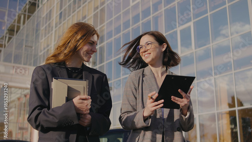 Two women walk and talk outside a modern office building in the late afternoon sun while carrying documents and a tablet