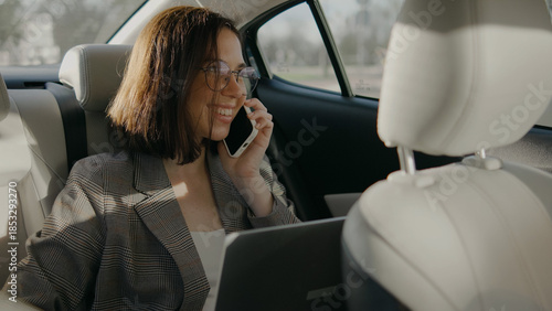 Woman talking on phone and working on laptop in back seat of car during daytime in a city environment