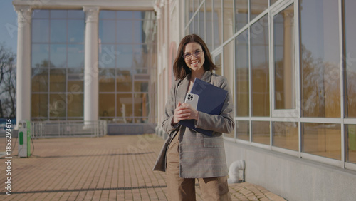Young woman stands outside a building holding a laptop and phone while smiling in bright sunlight during the day