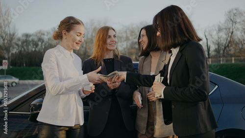 Women exchanging cash in a parking lot during an afternoon gathering while standing around a parked car