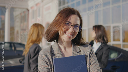 Group of women in business suits stand outside an office building during the day while one woman smiles and holds a briefcase