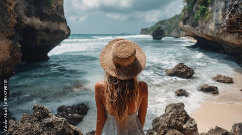 A woman stands on a beach looking out at the ocean. The scene includes rocky cliffs waves rolling in and a cloudy sky. It is daytime in Bali.